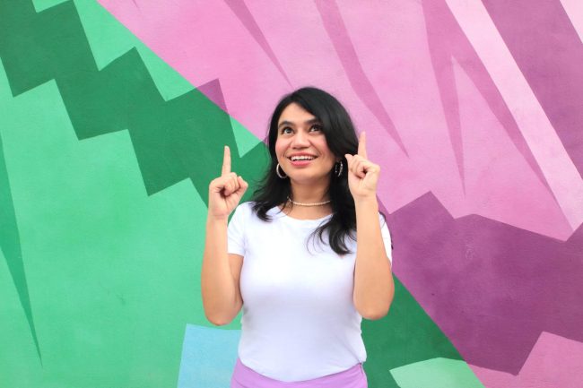 Judy Monsiváis copywriter y estratega Judy señala arriba con las dos manos con un fondo colorido